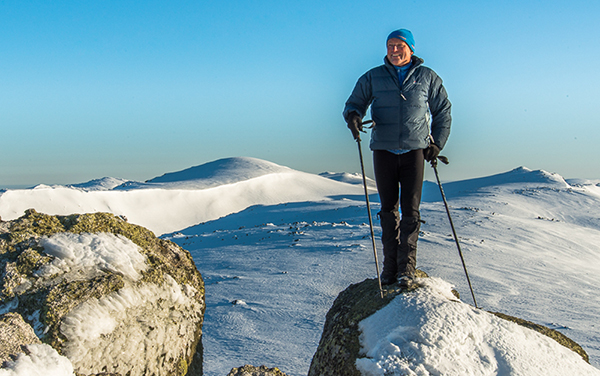 Photo of a man standing with walking poles on rock with snowy mountains behind him